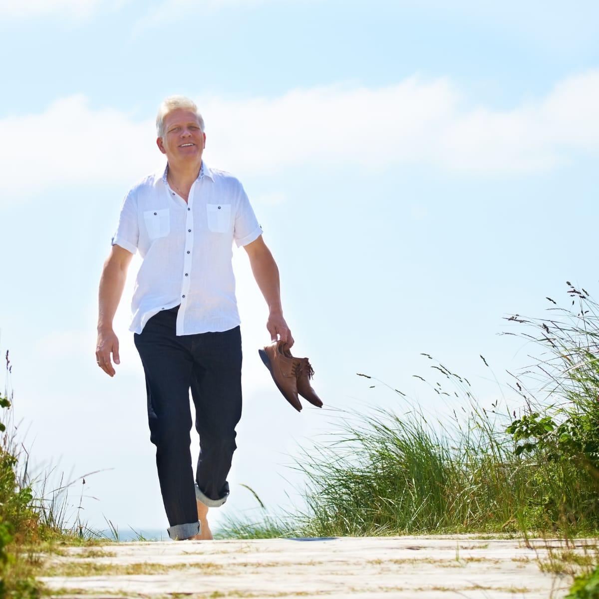 A smiling older man walking barefoot along a boardwalk by the beach, carrying his shoes in one hand