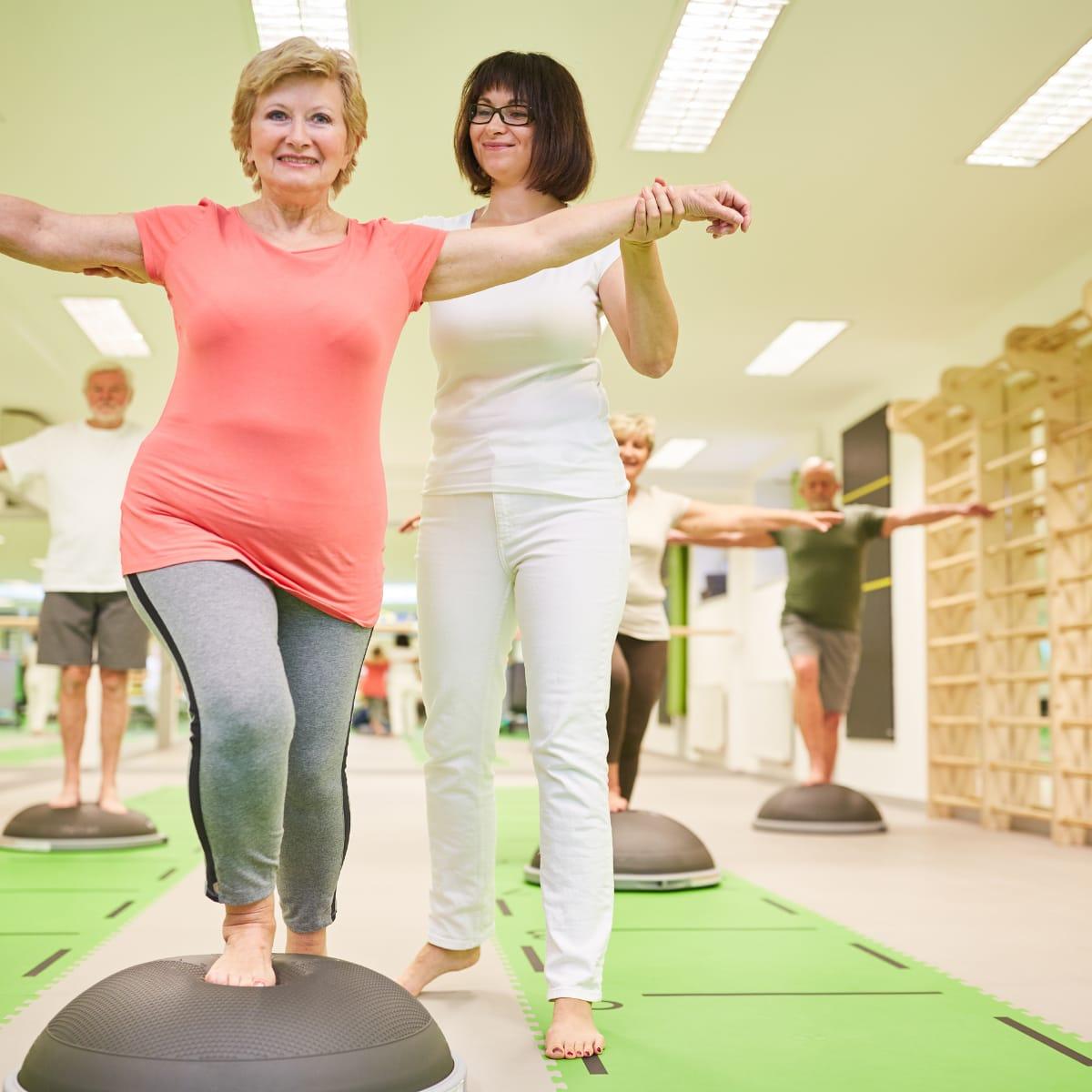 An older woman balancing on a half-dome trainer while a coach steadies her arm in a group class