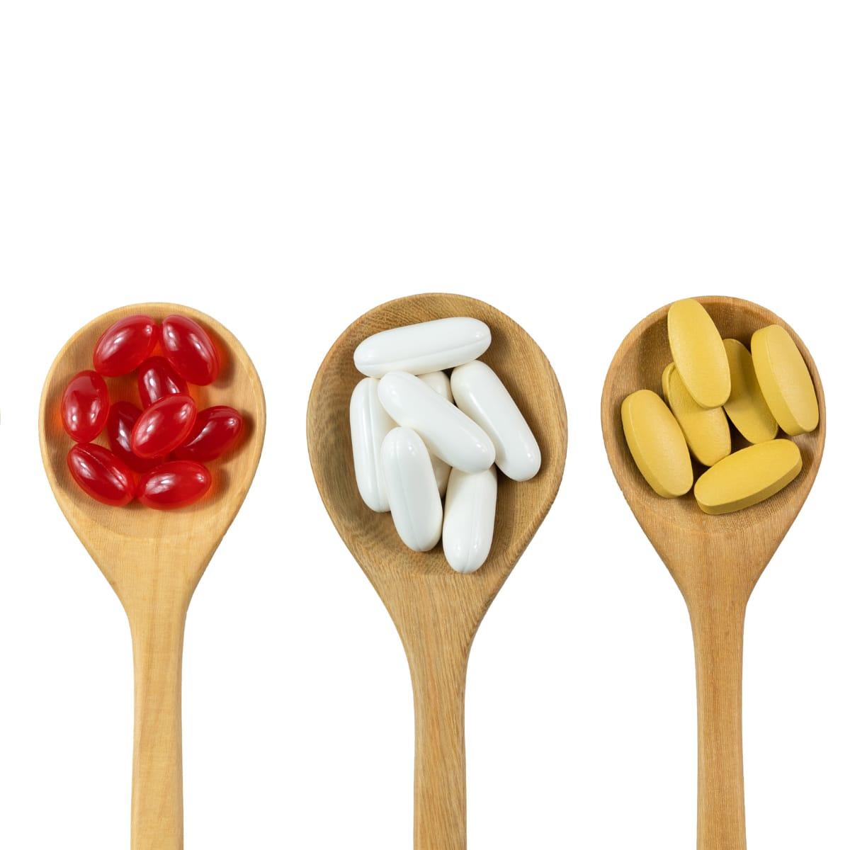 Three wooden spoons holding red softgels, white capsules and yellow tablets