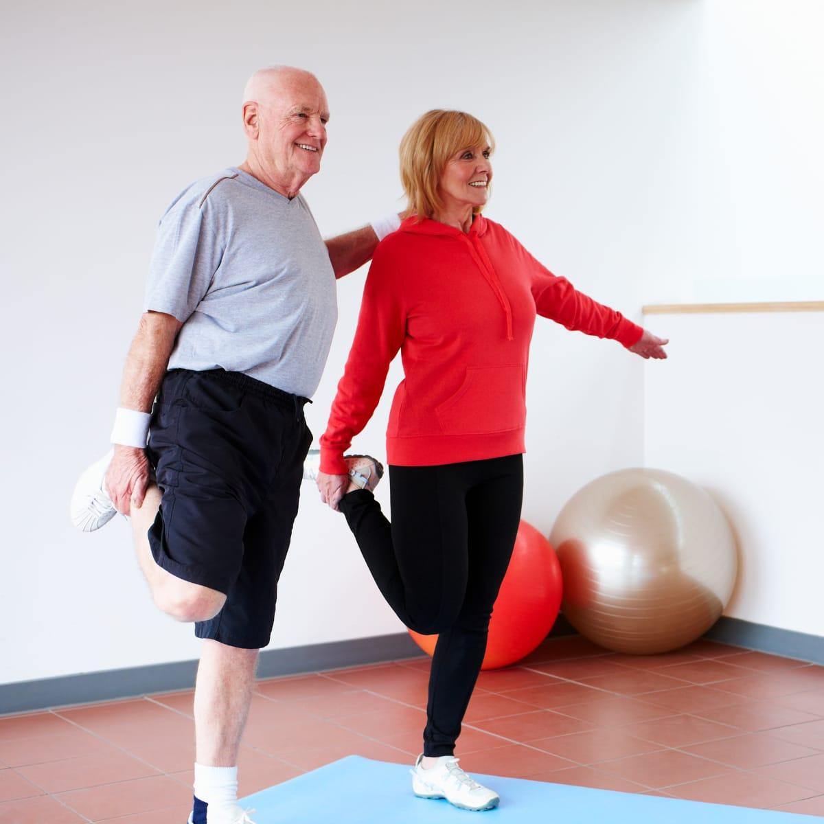 An older man and woman smiling as they balance on one leg in an exercise studio, each holding a foot behind them in a quad stretch