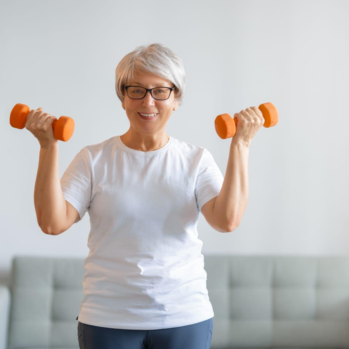 A smiling older woman in a white T-shirt doing bicep curls with orange dumbbells in her living room