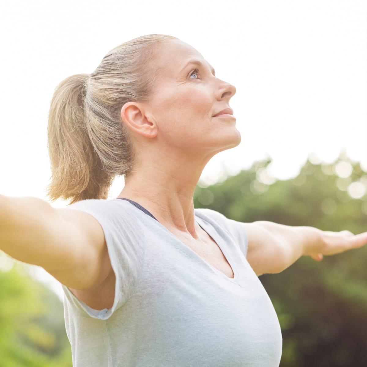 A woman with a ponytail standing outdoors with her arms stretched wide and face turned up to the sun