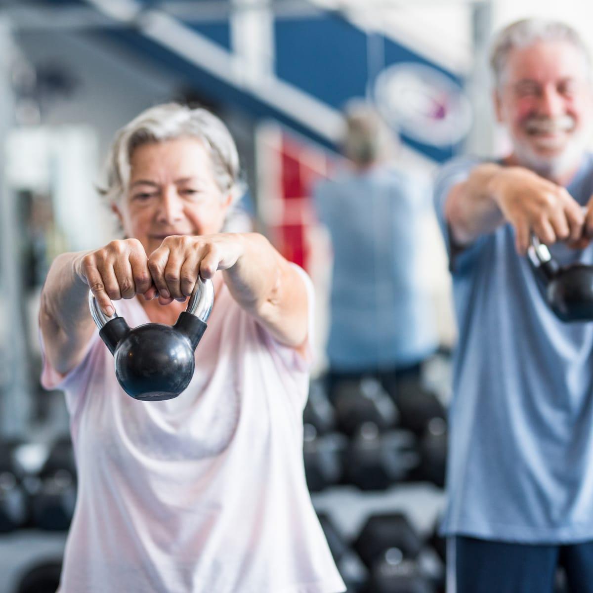 An older woman swinging a kettlebell in a gym with an older man training behind her