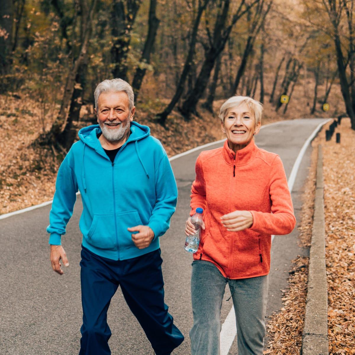 A smiling older couple walking briskly together on a tree-lined road in autumn