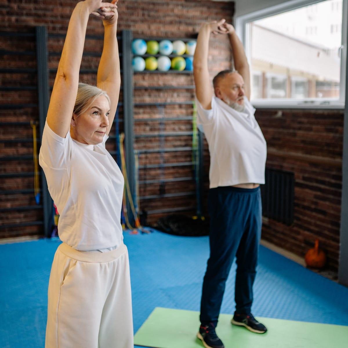 An older woman and man reaching both arms overhead in an exercise studio