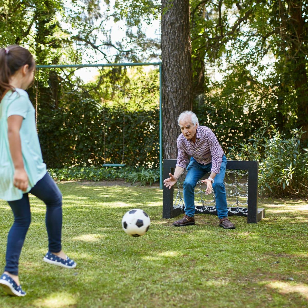 A grandfather crouching in front of a small backyard goal as his granddaughter kicks a soccer ball
