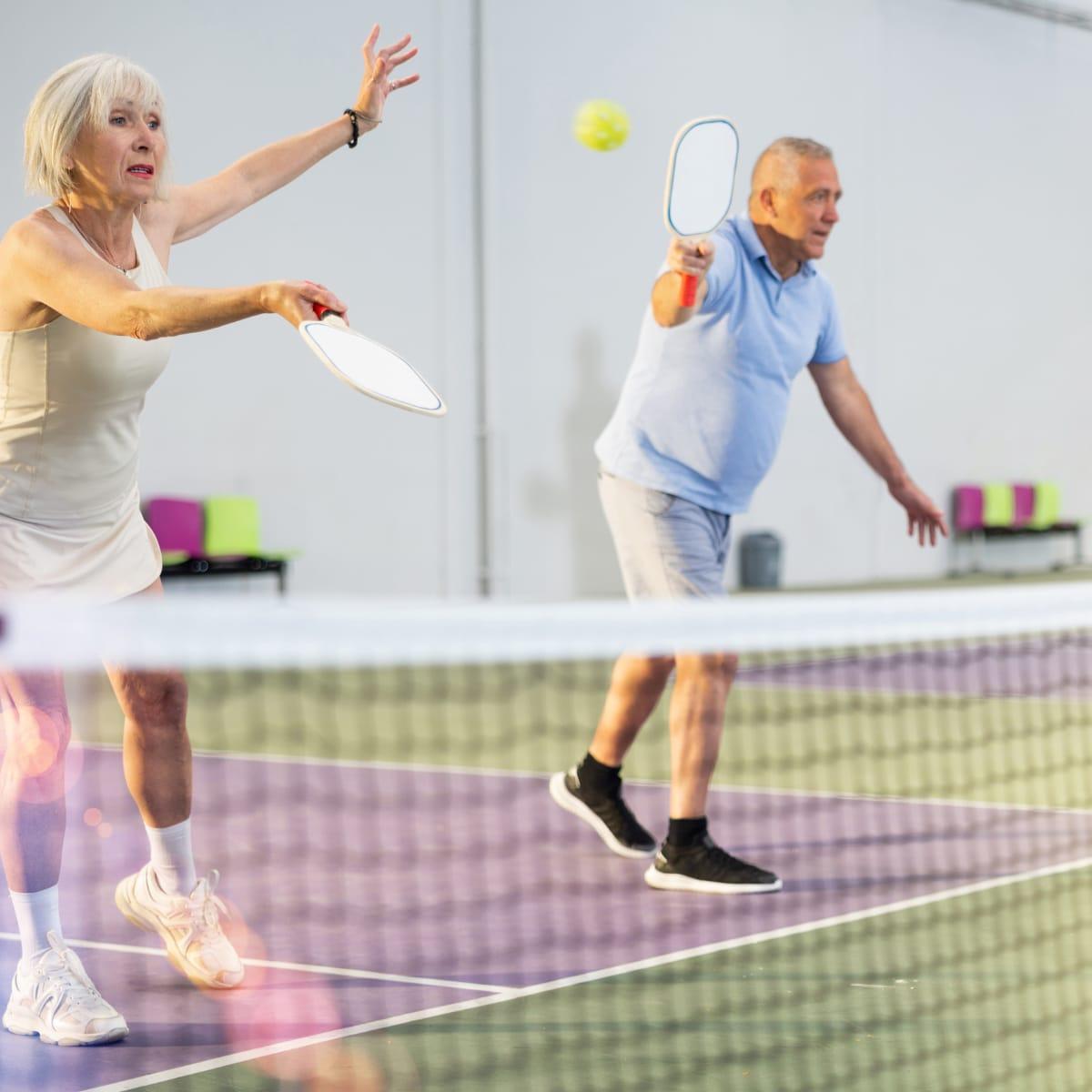 An older woman and man playing pickleball at the net on an indoor court