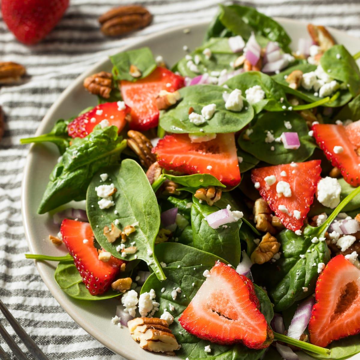 A plate of spinach salad with sliced strawberries, crumbled cheese, red onion and nuts