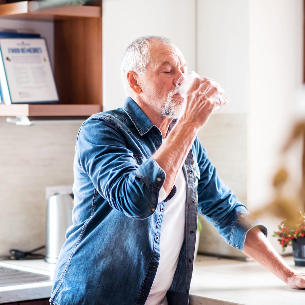 An older man with a gray beard drinking a glass of water in his kitchen
