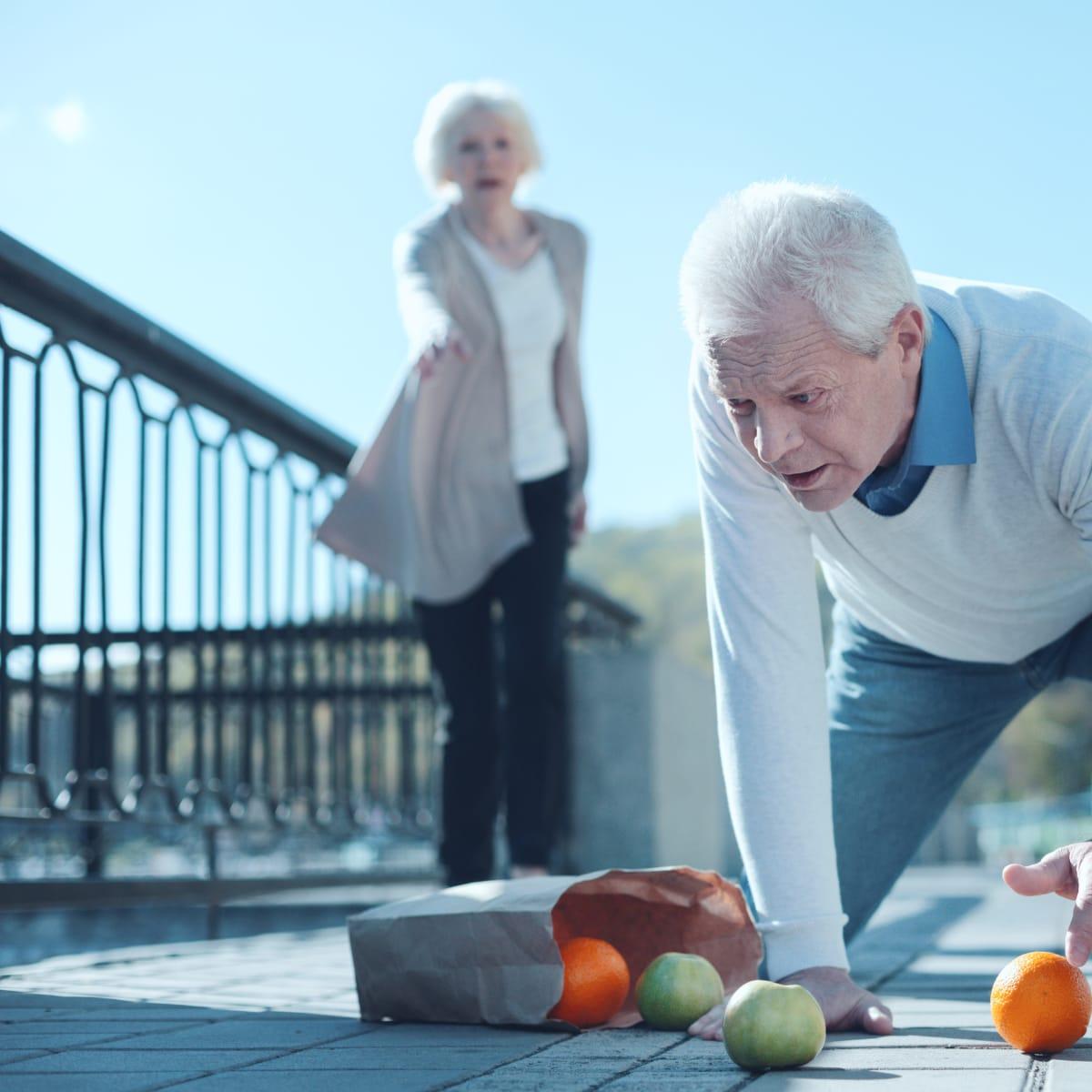 An older man catching himself on the pavement after a fall, with oranges and apples spilling from a paper bag while a woman reaches toward him