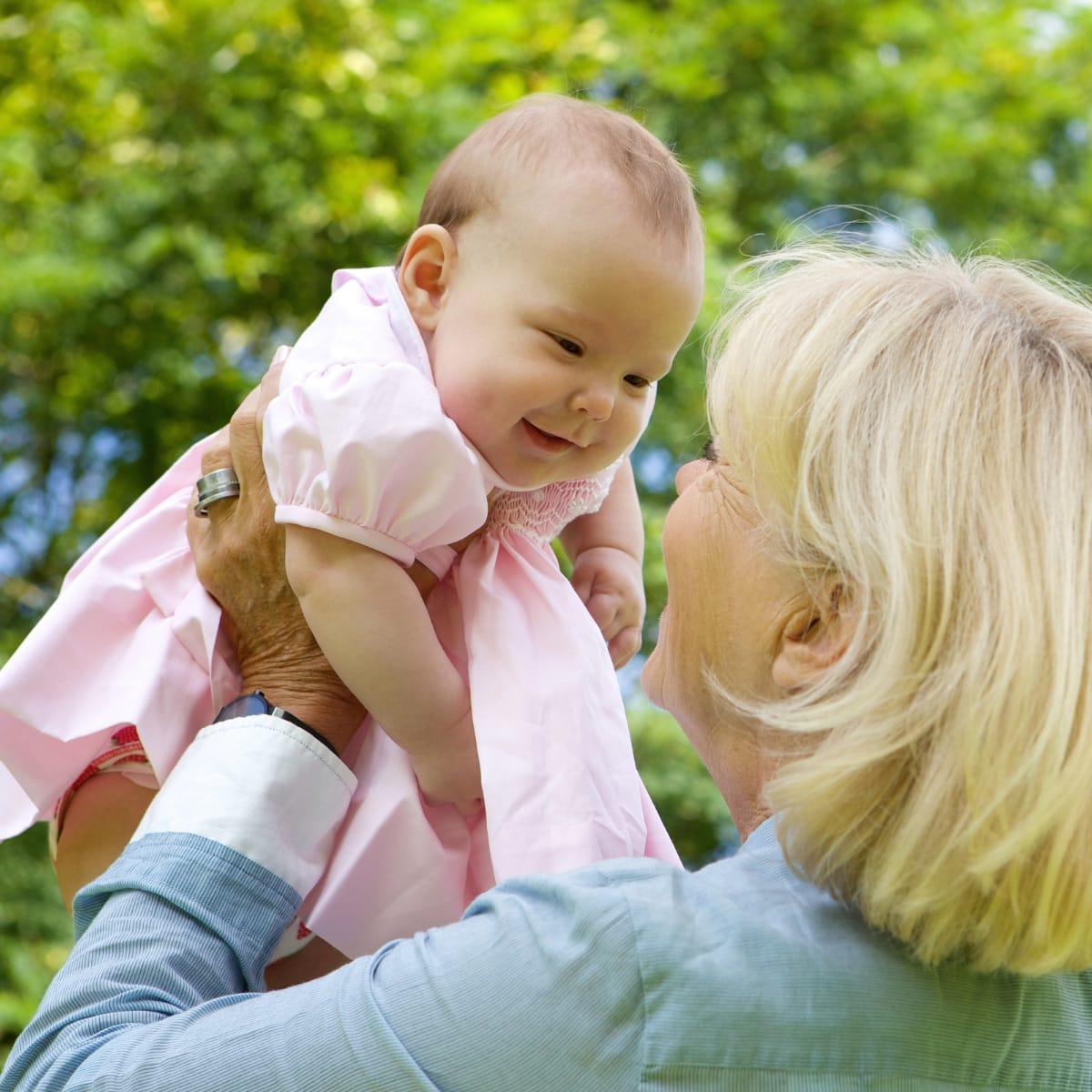 A smiling grandmother lifting a laughing baby in a pink dress up into the air outdoors