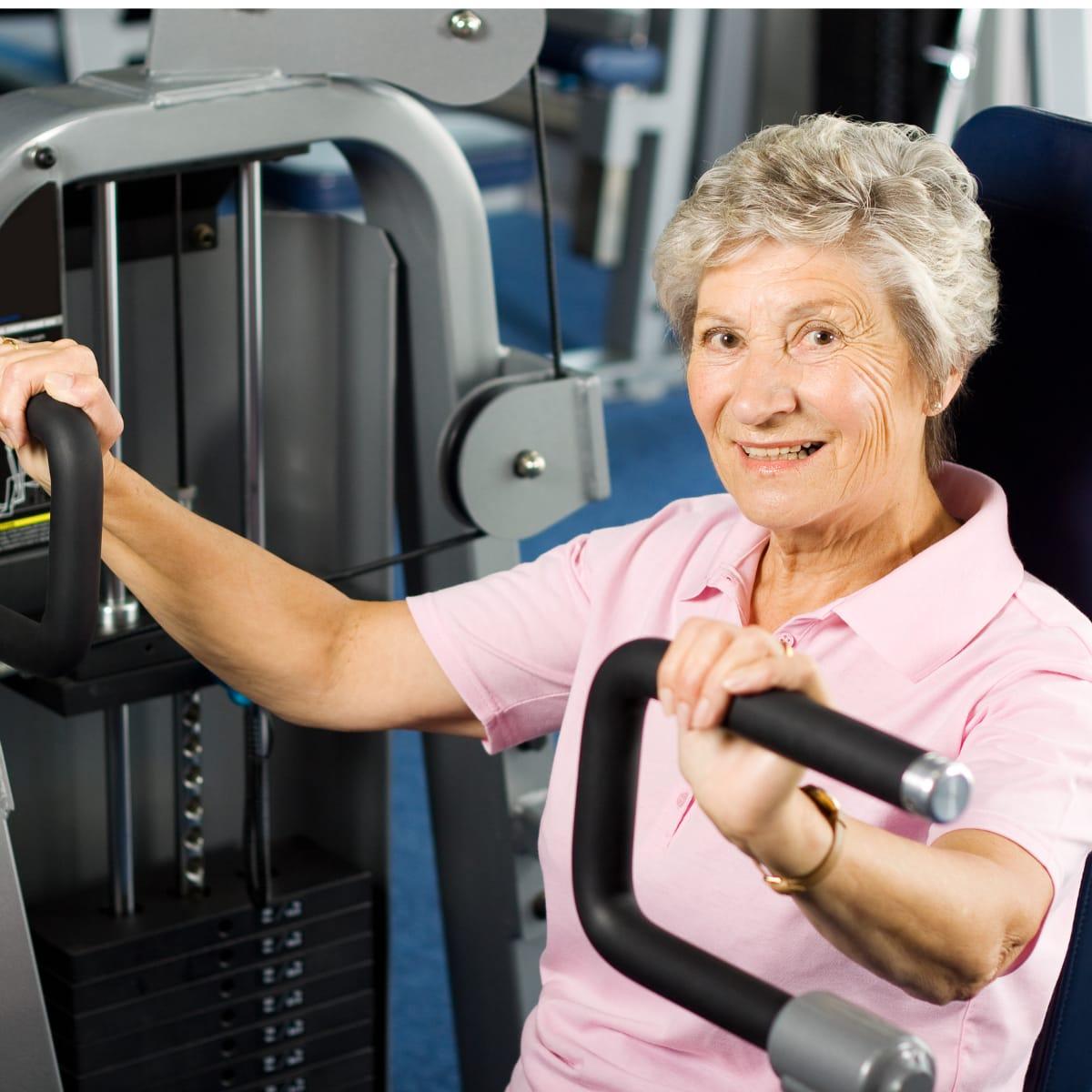 A smiling older woman in a pink polo shirt using a chest press machine at the gym