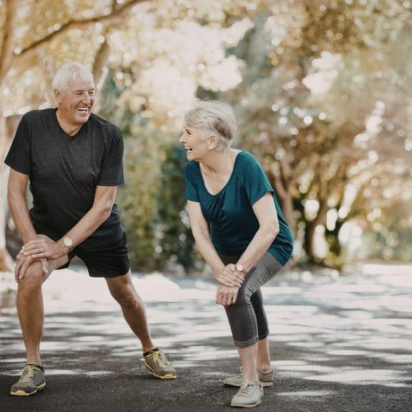 An older man and woman laughing as they stretch their legs together on a tree-lined path