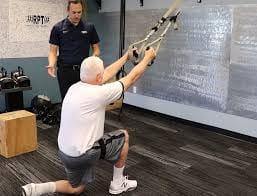An older man kneeling and pulling on suspension straps while a trainer watches in a gym
