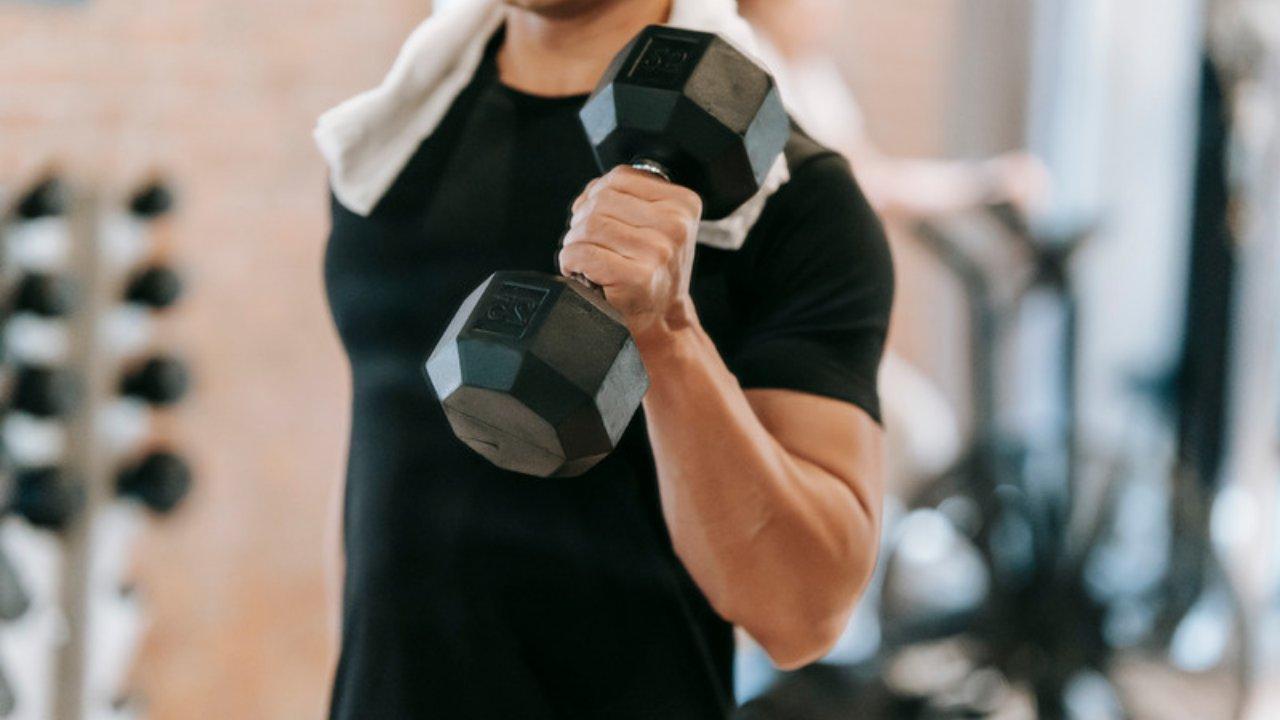 A man in a black T-shirt with a towel around his neck curling a dumbbell in a gym