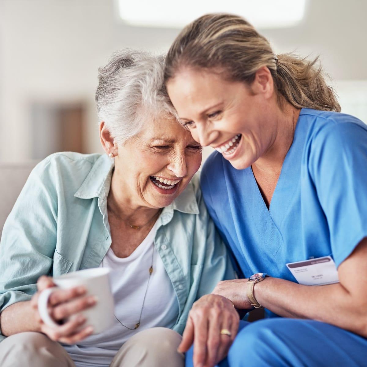 An older woman holding a mug and laughing with a caregiver in blue scrubs who leans in close beside her