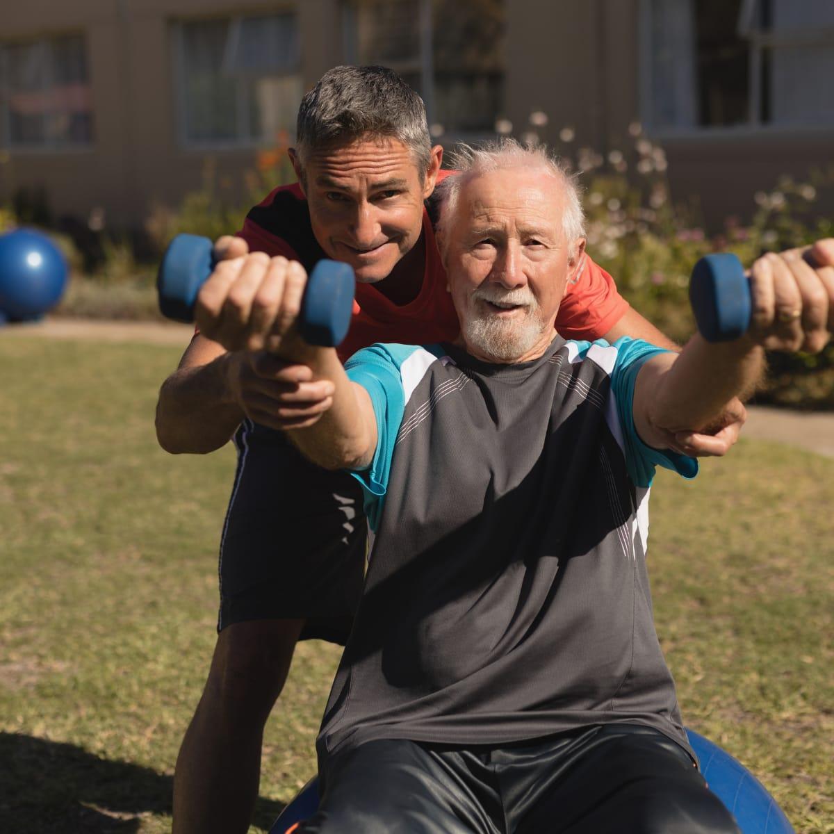 A trainer standing behind a smiling older man, supporting his arms as he lifts dumbbells outdoors