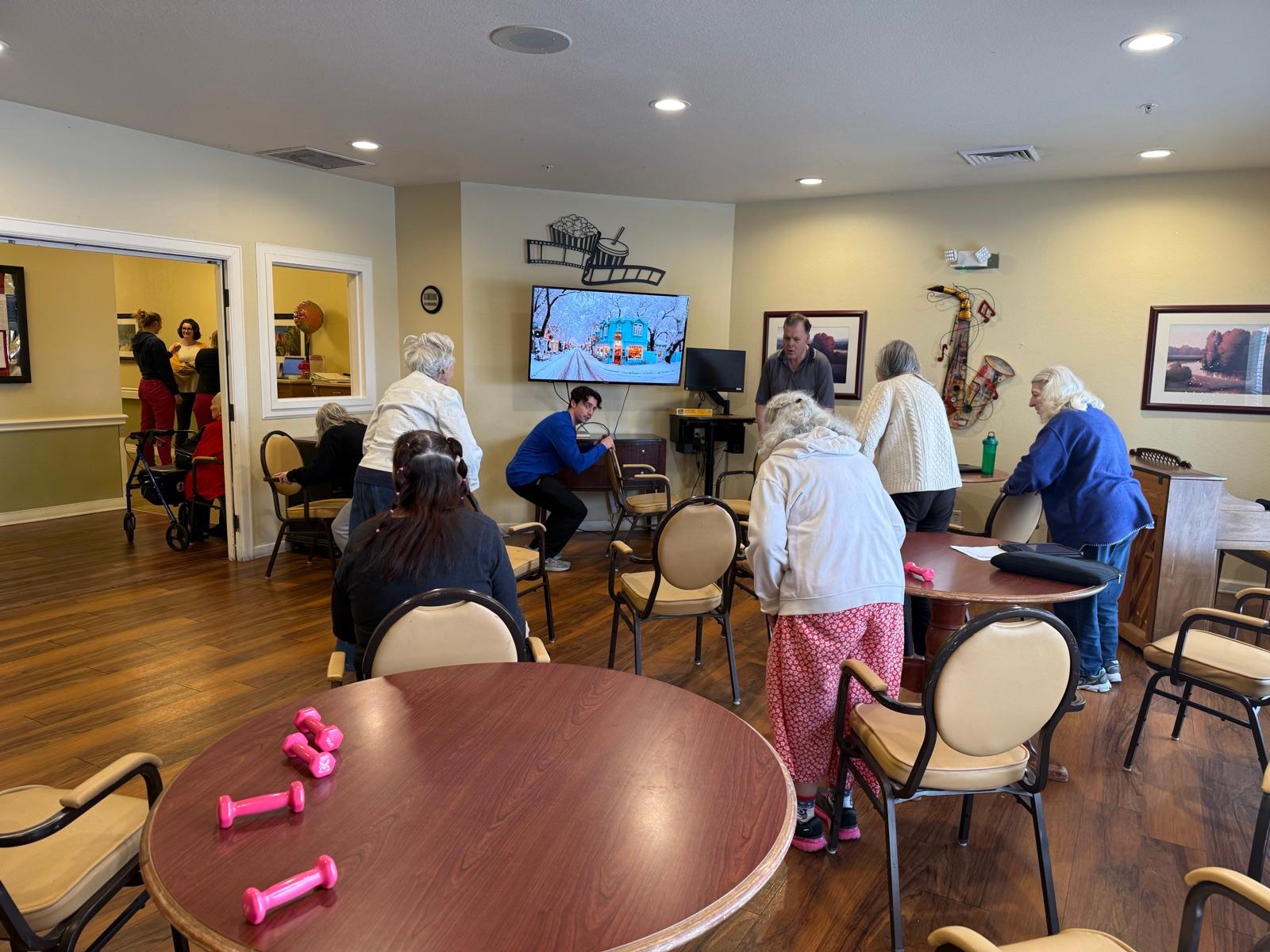Residents following Alex through a chair squat during a TogetherWON class, with pink hand weights on the table
