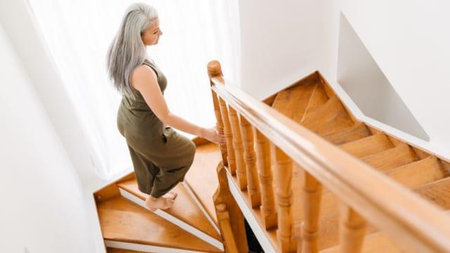 A gray-haired woman climbing a wooden staircase at home, holding the handrail