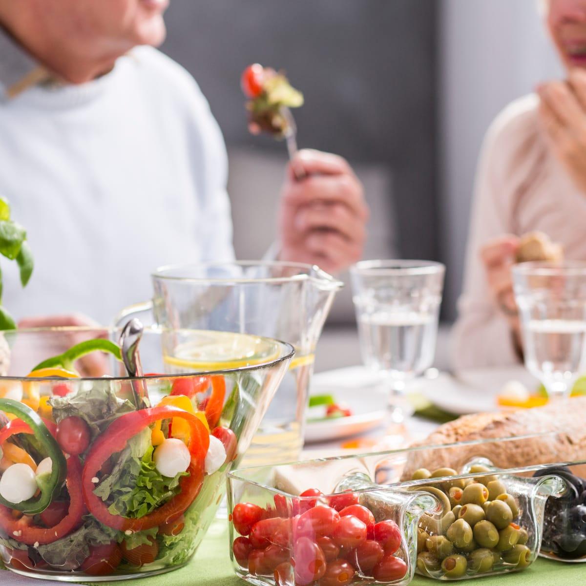 An older couple sharing a meal with a fresh salad, cherry tomatoes and olives on the table