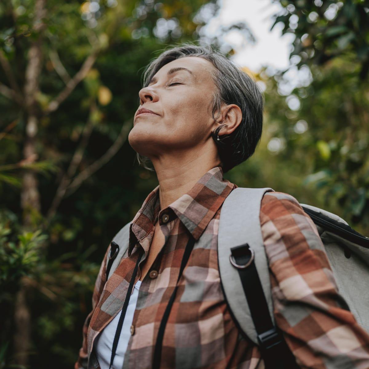 A woman with a backpack pausing on a forest trail, eyes closed, taking a deep breath