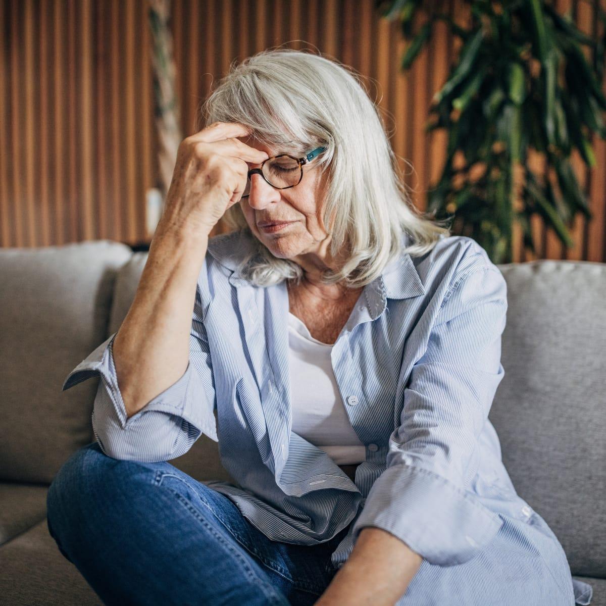 A stressed older woman with glasses sitting on a sofa, eyes closed, hand pressed to her forehead