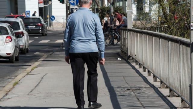 An older man in a blue shirt walking away along a city sidewalk beside parked cars