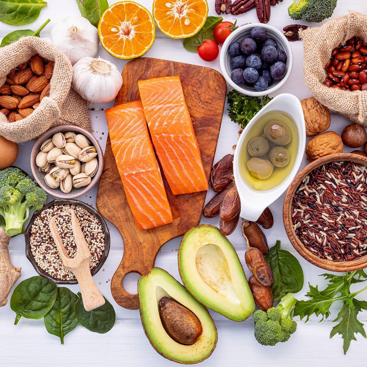 Healthy whole foods on a white table, including salmon fillets, avocado, nuts, quinoa, wild rice, olive oil, blueberries and broccoli