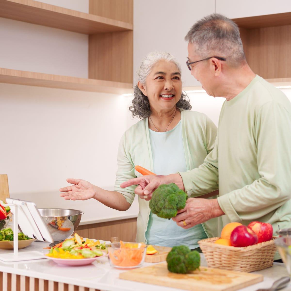 A smiling older couple preparing vegetables together in a bright kitchen, the man holding a head of broccoli