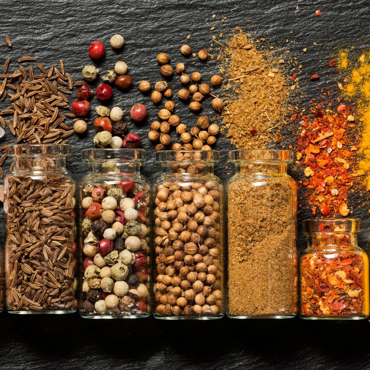 A row of glass jars filled with colorful whole and ground spices on dark slate