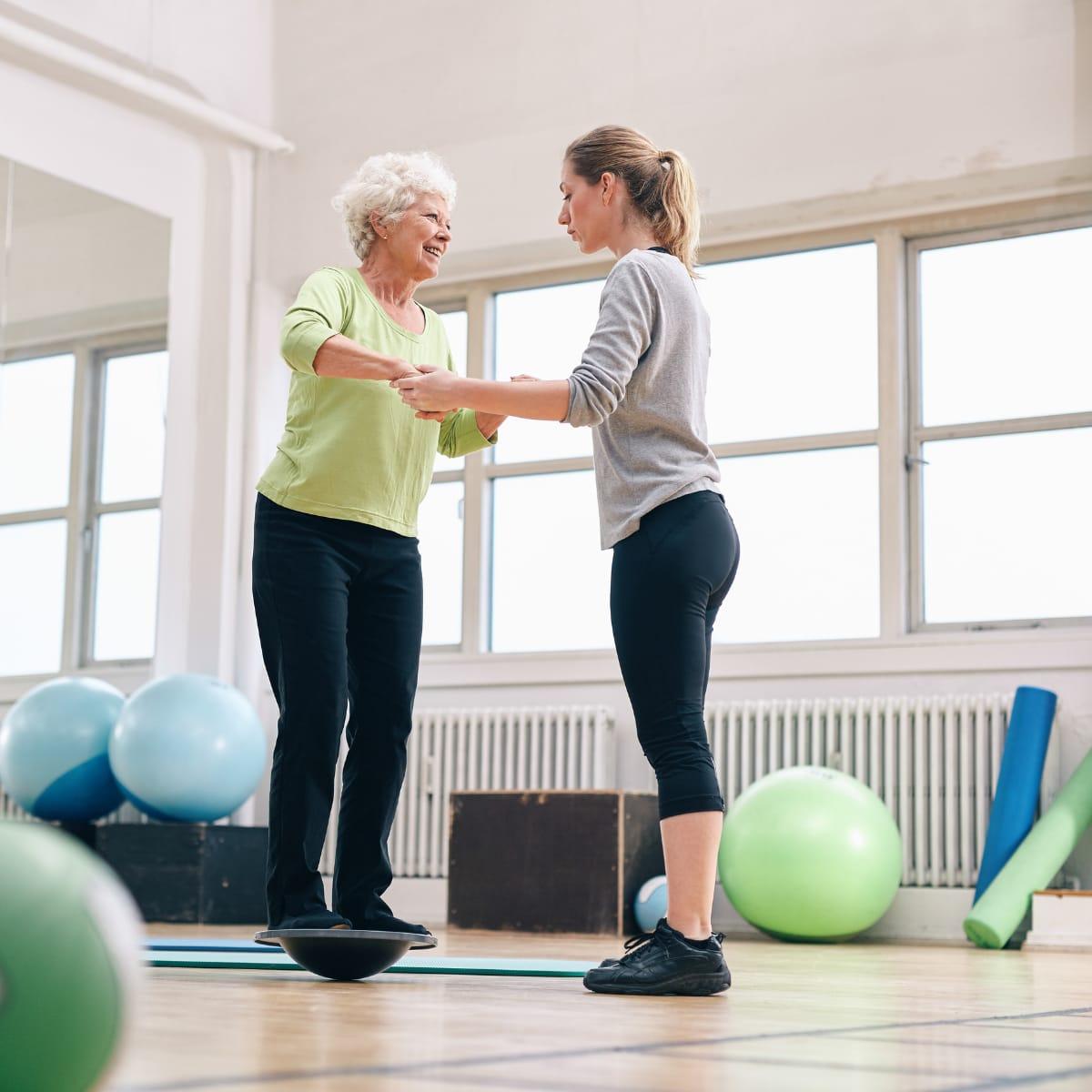 A trainer holds the hands of a smiling older woman balancing on a wobble board