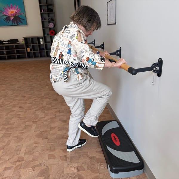 Brenda holding a wall-mounted handrail as she lifts one foot onto an exercise step