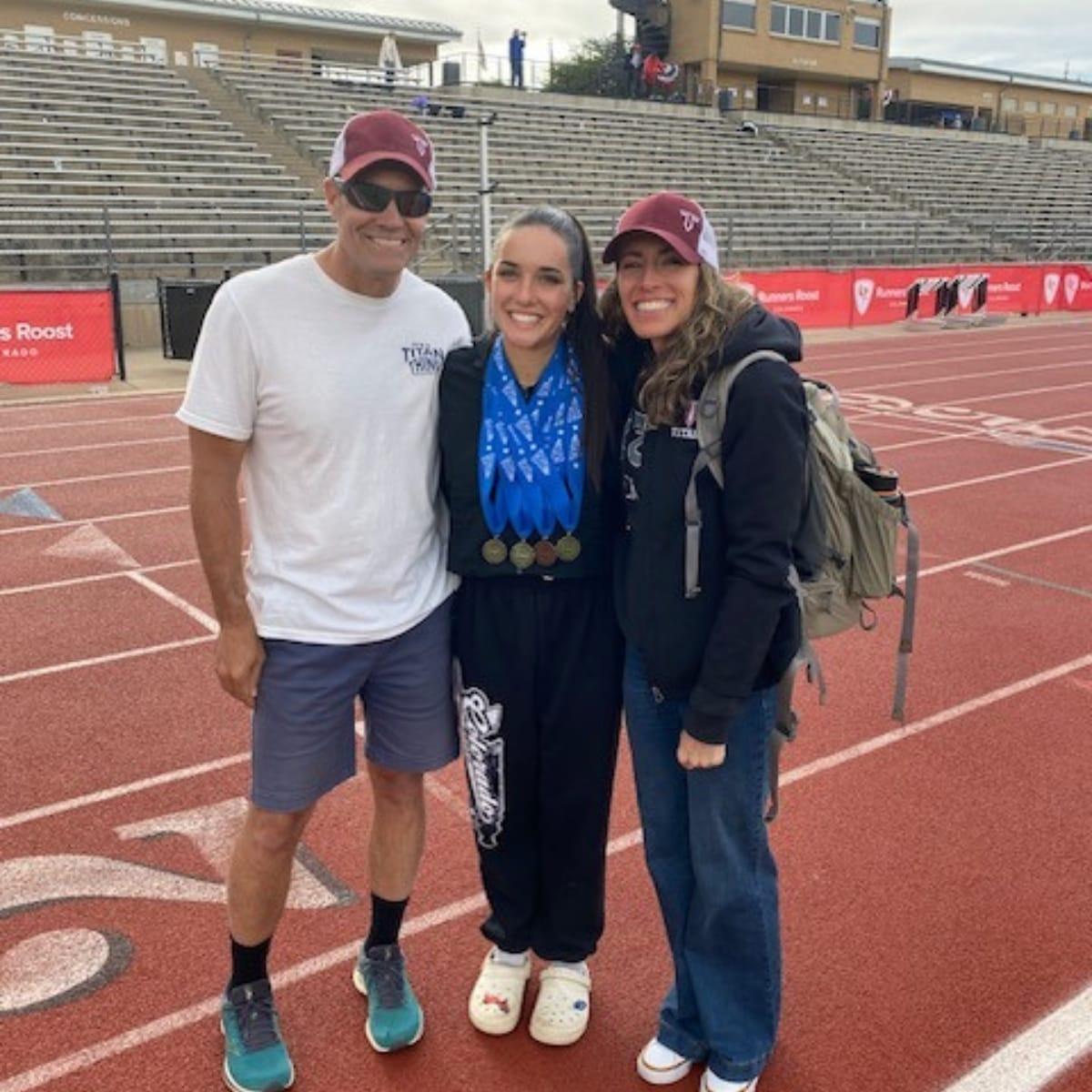 Aaron standing on a red running track with his two daughters, one wearing several medals