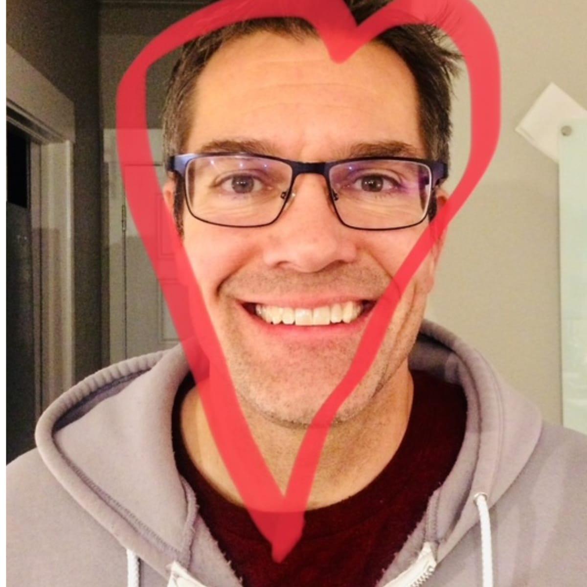A smiling Aaron in glasses and a gray hoodie, with a red heart drawn around his face