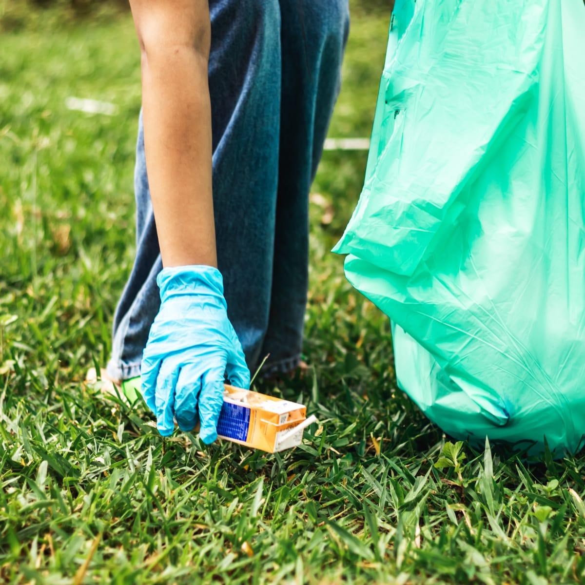 A gloved hand bending down to pick up litter from the grass beside a green trash bag