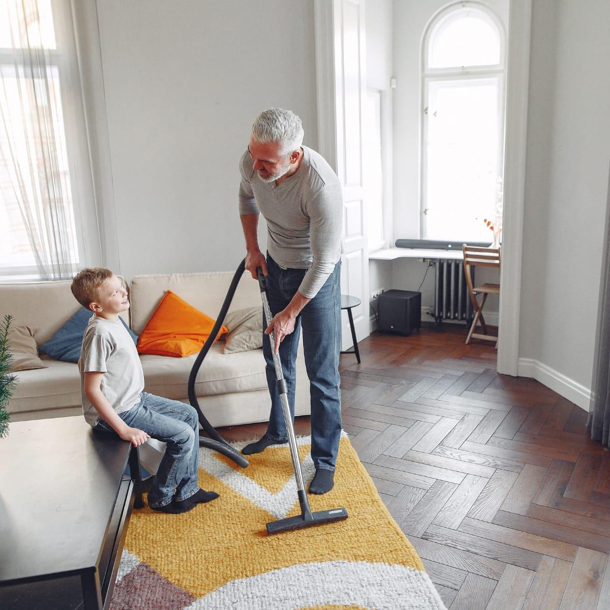 A grandfather vacuuming a rug while his grandson watches from the coffee table