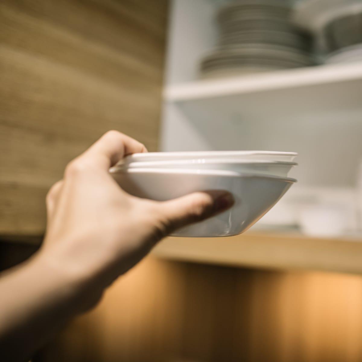 A hand reaching to place a stack of white bowls on a kitchen cabinet shelf