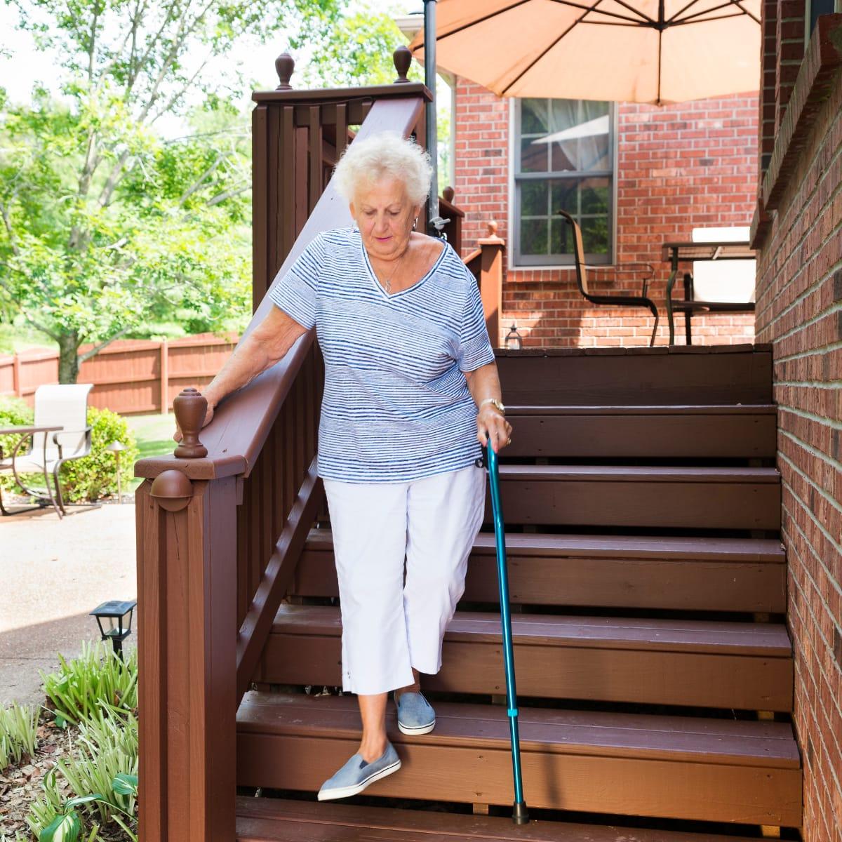 An older woman with a cane walking down her deck steps while holding the railing