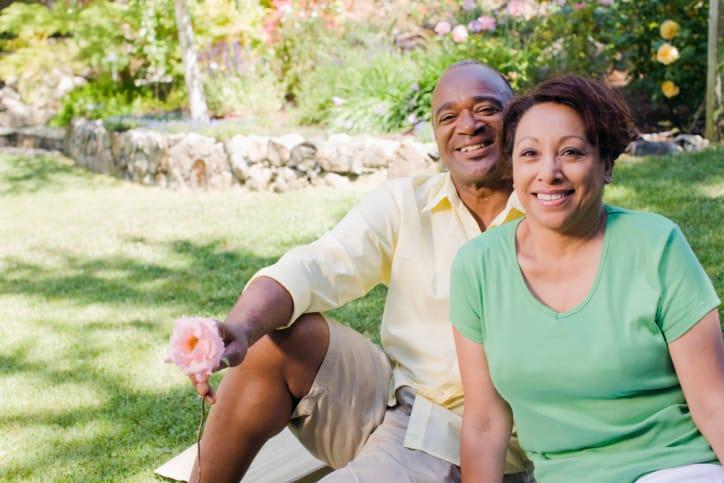 A smiling older couple sitting on the grass in a flower garden, the man holding a pink rose