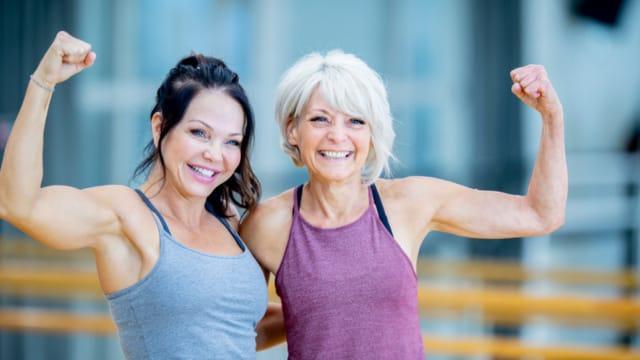 Two smiling women in tank tops flexing their arms in a fitness studio