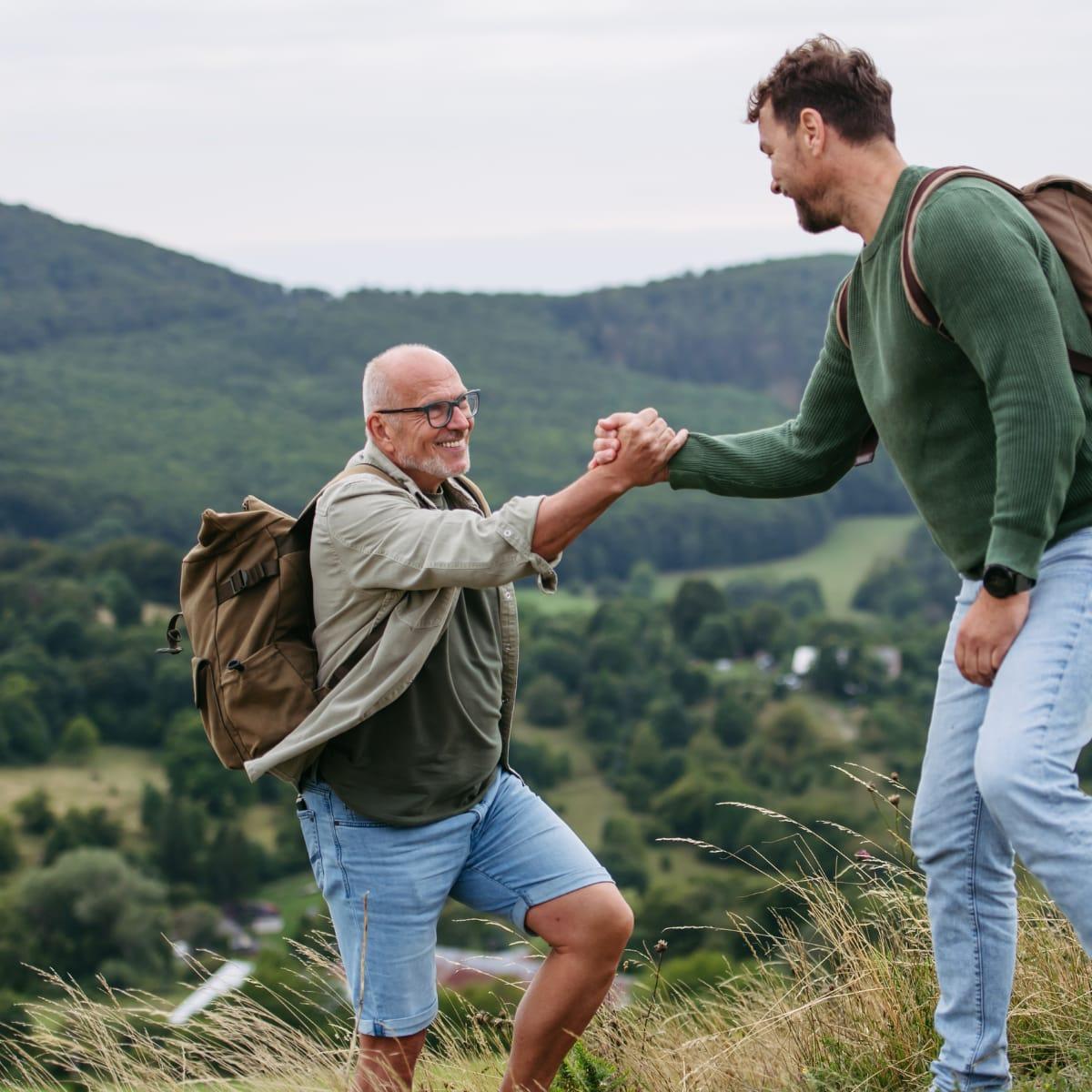 An older hiker with a backpack taking a friend's hand to step up a grassy hillside above a green valley