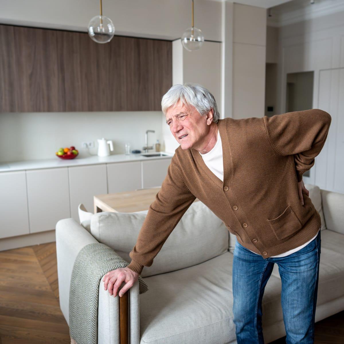 An older man grimacing and holding his lower back as he pushes himself up from a sofa