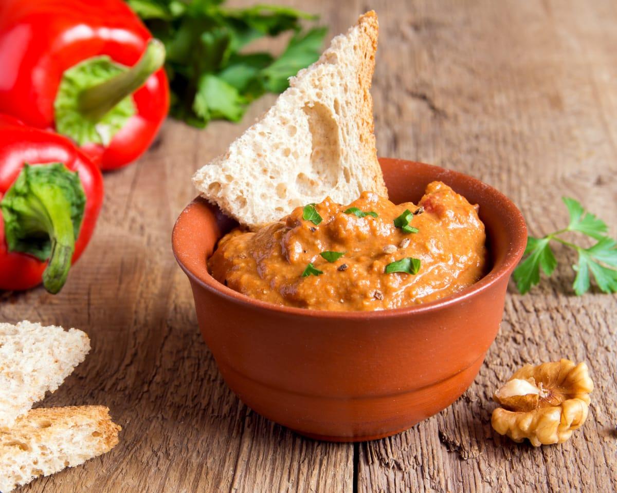A terracotta bowl of orange red pepper dip with a piece of bread, beside fresh red peppers on a wooden table
