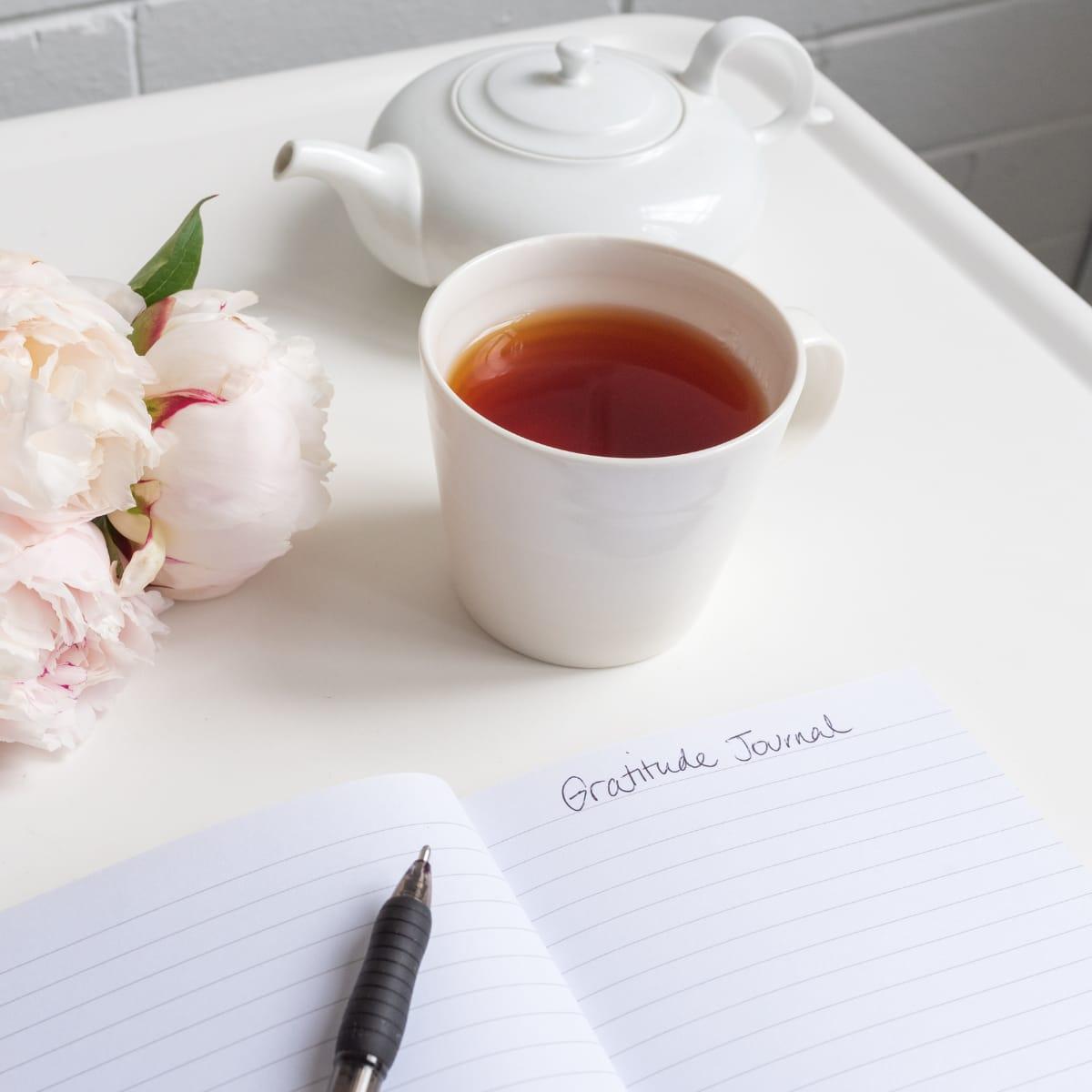 An open notebook titled "Gratitude Journal" with a pen, beside a cup of tea and pink peonies