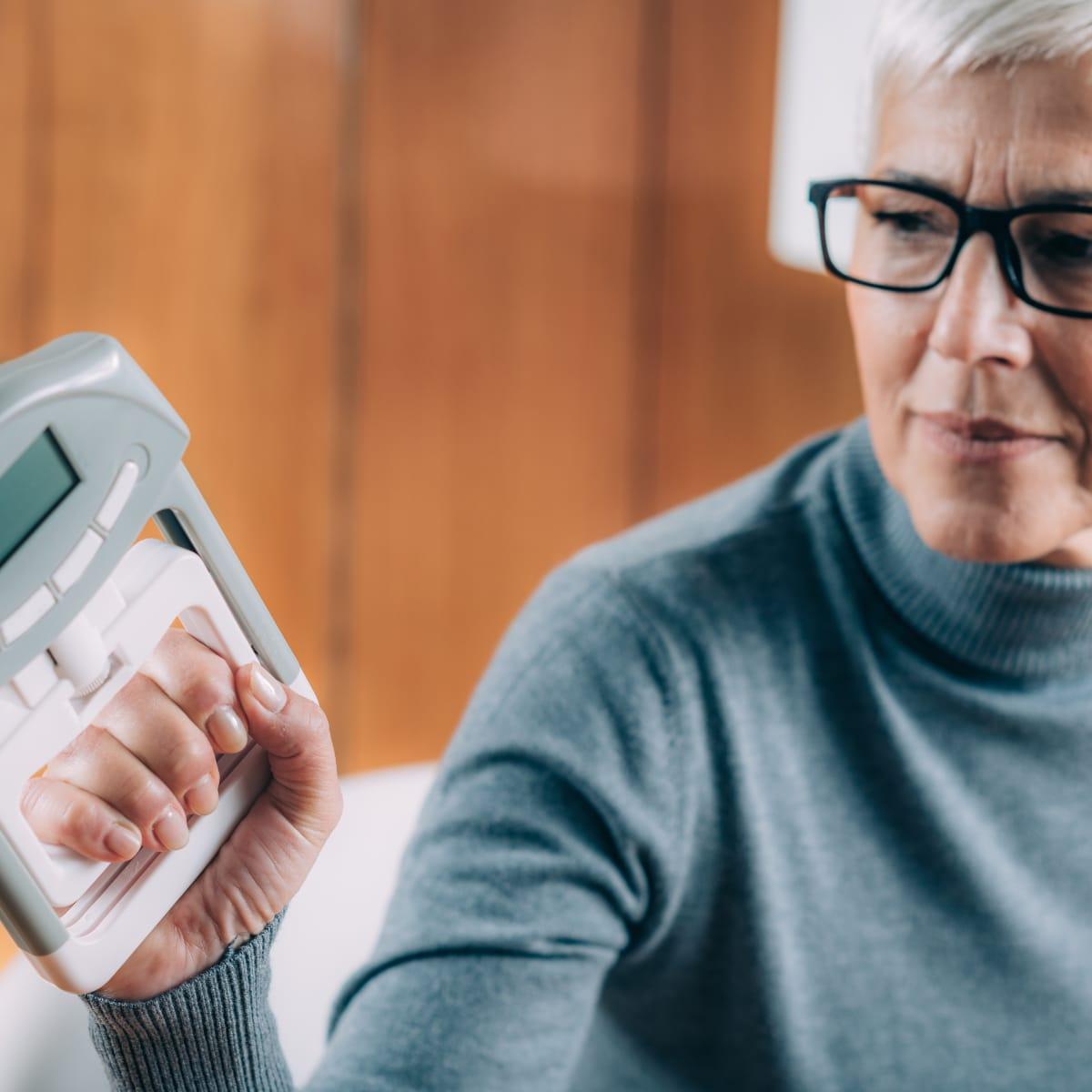 An older woman in glasses squeezing a digital hand grip dynamometer