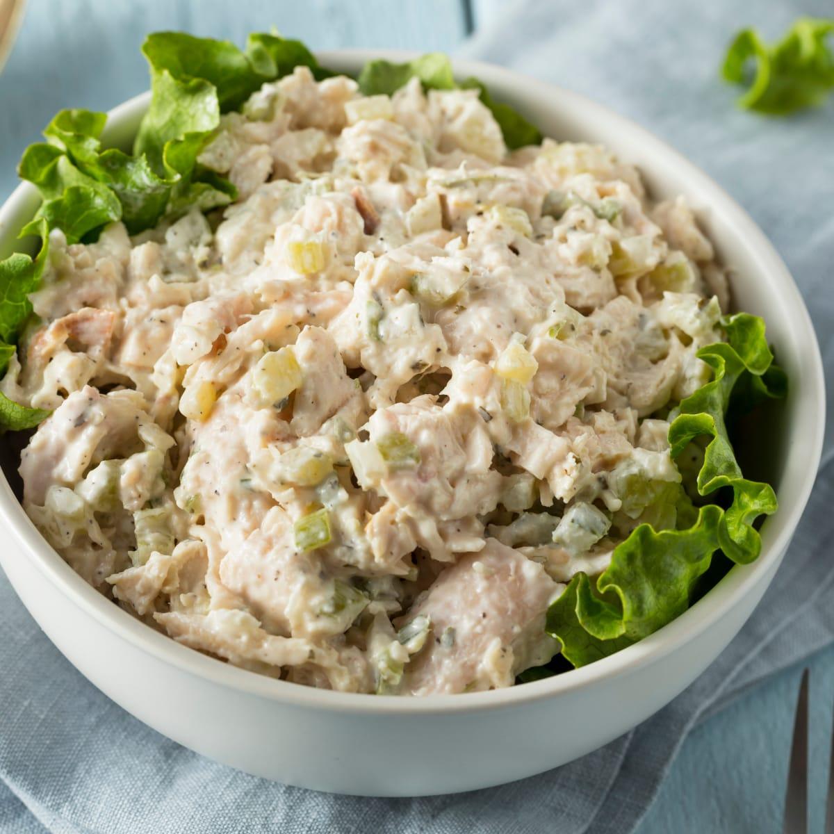 A white bowl of creamy chicken salad with diced pickles and celery on lettuce leaves