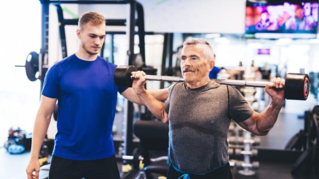 An older man doing a barbell squat in a gym while a young trainer watches