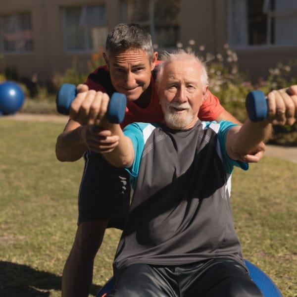 A trainer coaching an older man through a dumbbell exercise outdoors on the grass