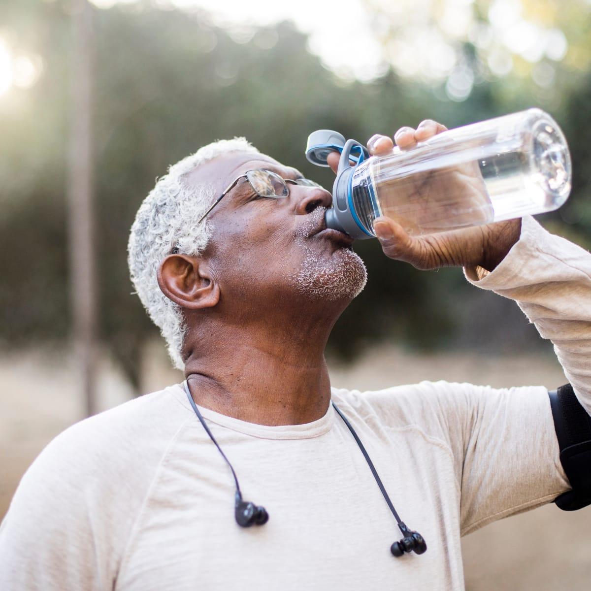 An older man with white hair and glasses drinking from a water bottle outdoors after exercise