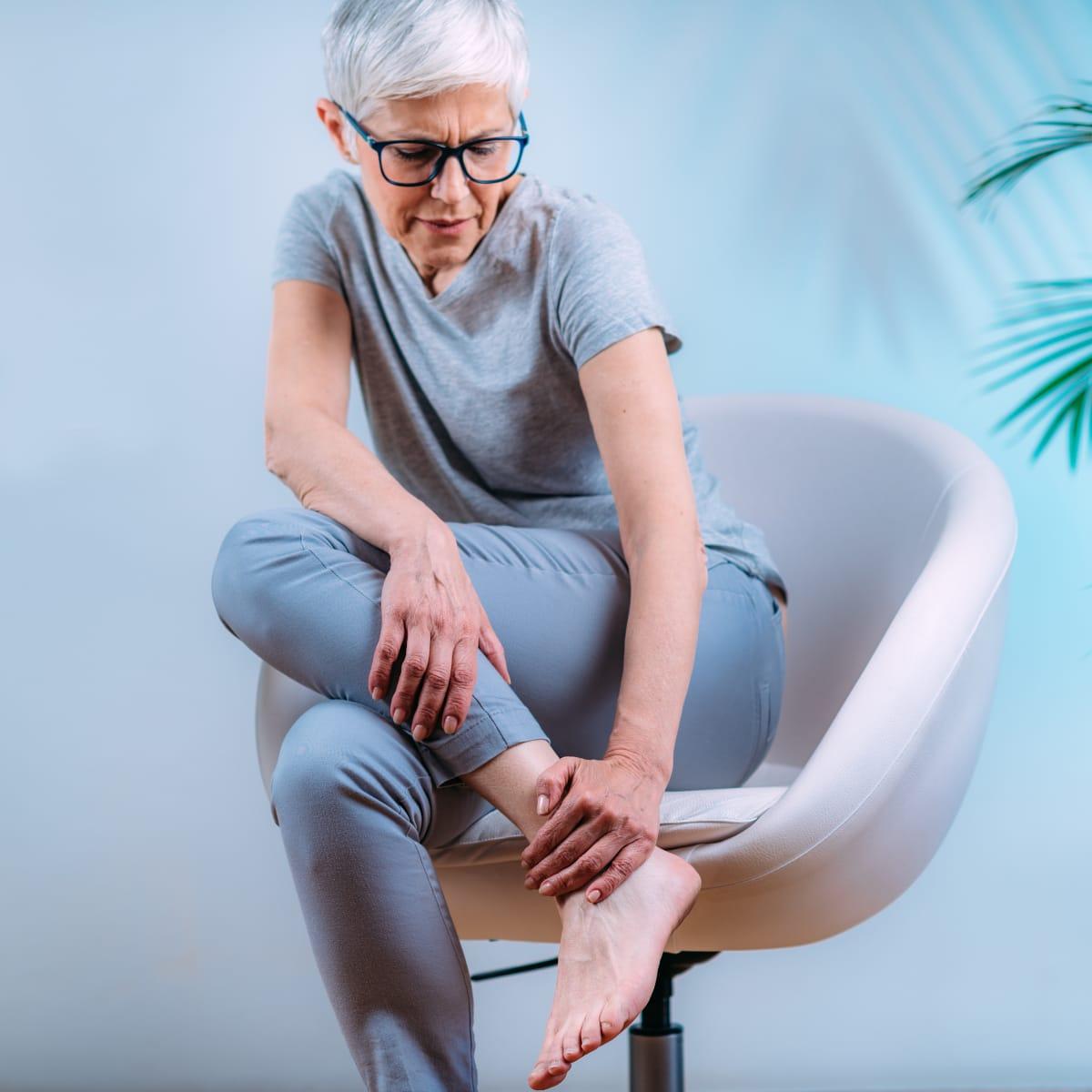An older woman with short gray hair and glasses sitting in a chair and holding her bare ankle as if it aches